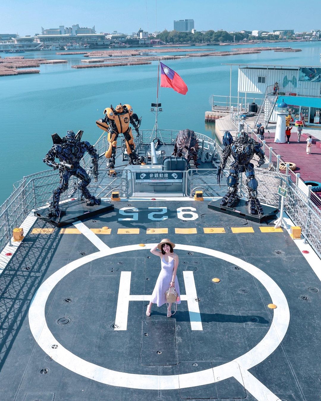 A woman in a white dress and hat stands on a ship's helipad, surrounded by large robot statues