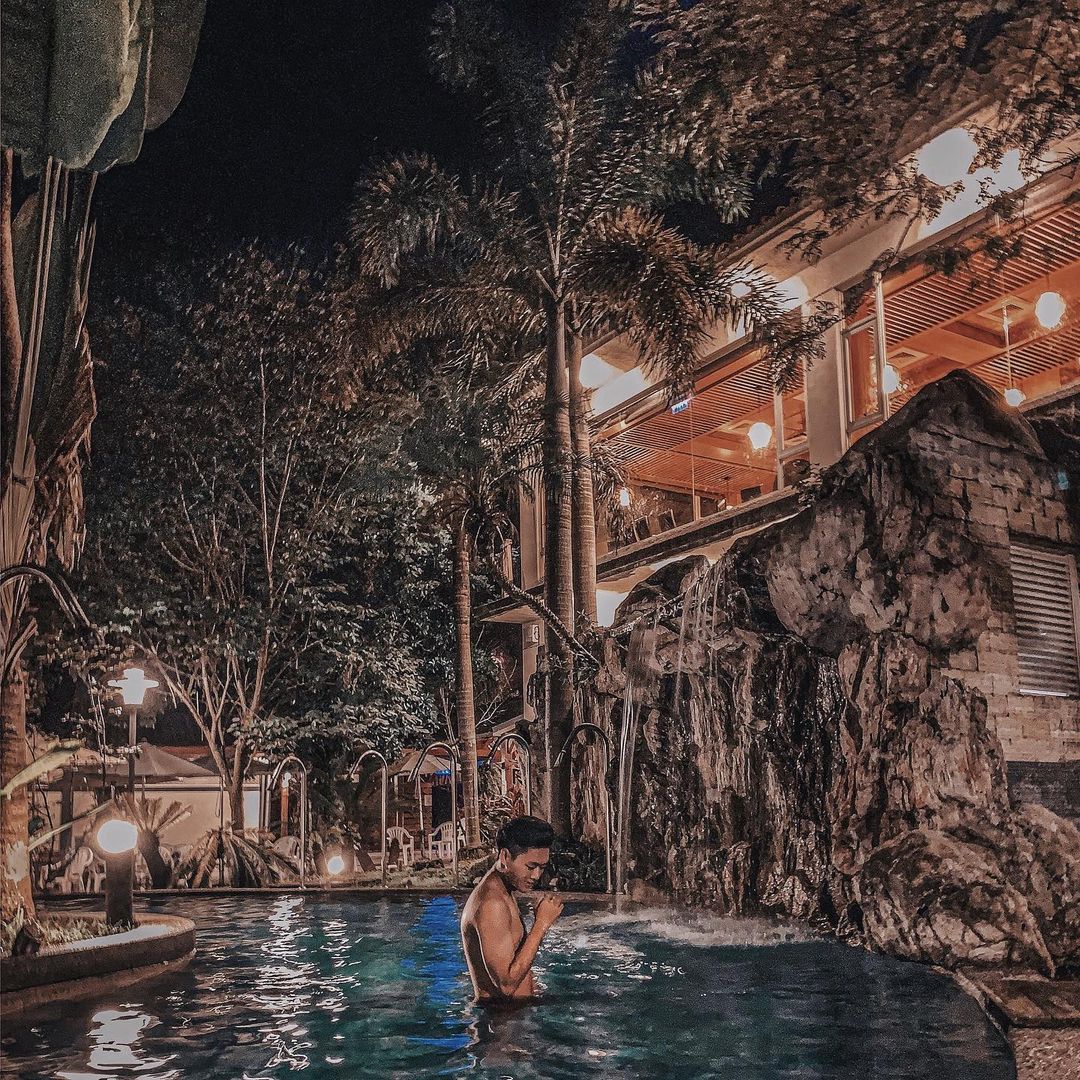 A man in a tropical pool at night with warm lighting and a stone waterfall