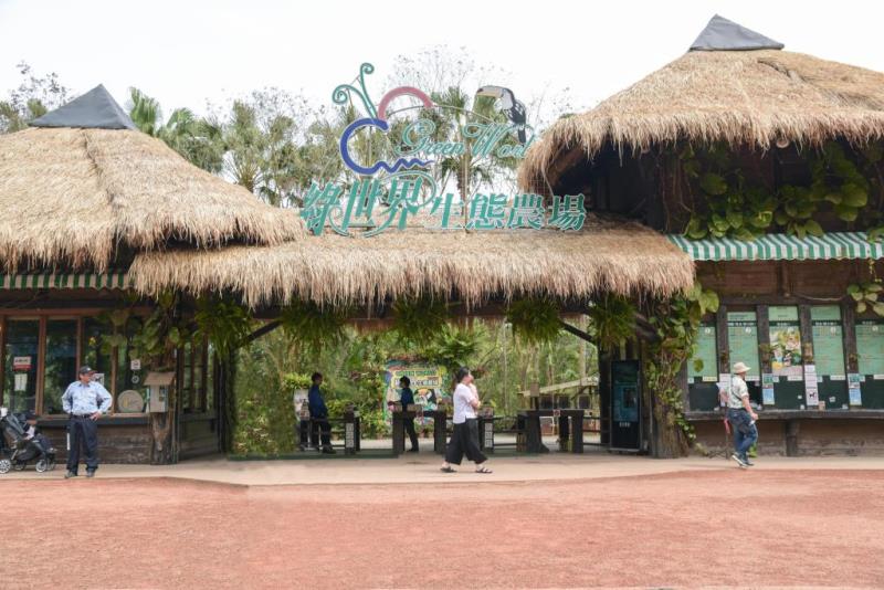 Entrance of a tropical-themed park with thatched roofs, lush greenery, and people walking