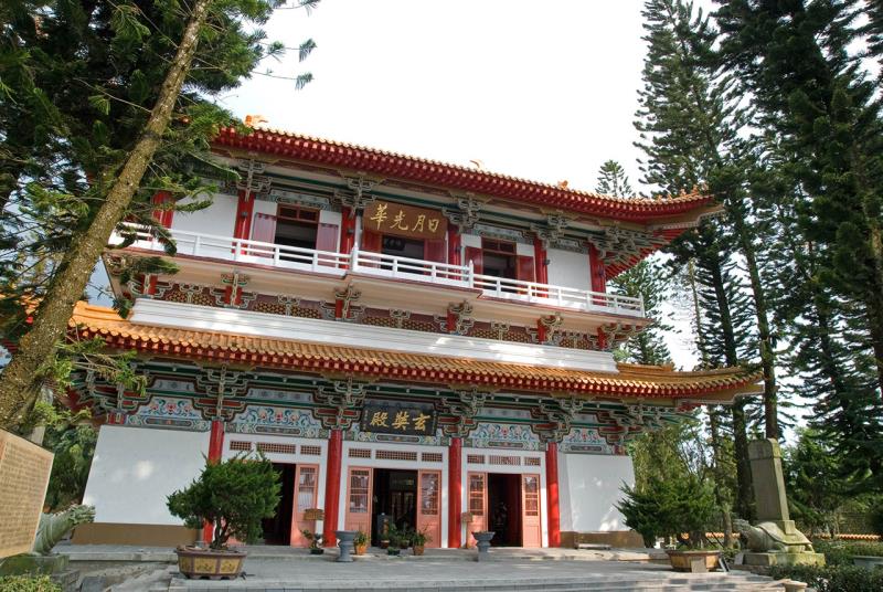 A traditional two-story Chinese temple with red and white detailing, surrounded by tall trees