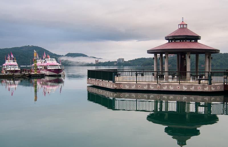 lakeside scene with a gazebo reflecting in water, two colorful boats docked nearby